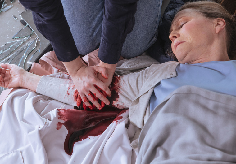 En kvinna med kraftigt s&aring;r p&aring; ena armen ligger p&aring; golvet medan tv&aring; h&auml;nder trycker p&aring; bl&ouml;dningen.